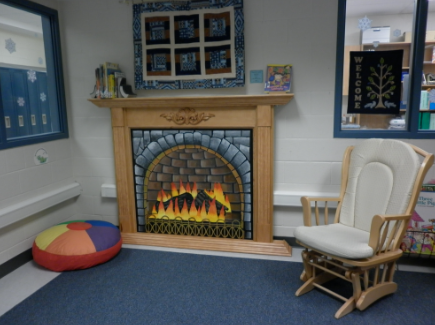corner of the library. A rocking chair is beside a pretend fireplace and a circular pillow with rainbow colours on it